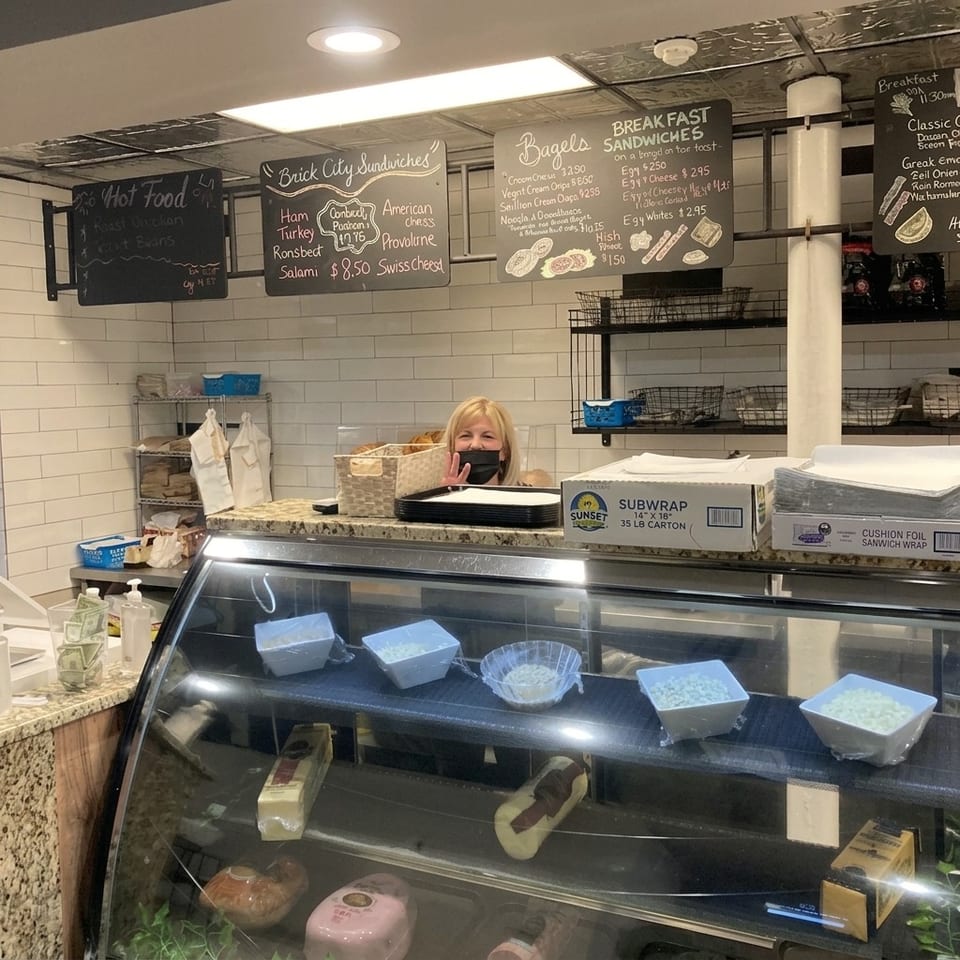 Sandwich shop counter with a masked employee, deli display case, ingredient bins, and chalkboard menus for sandwiches and breakfast.