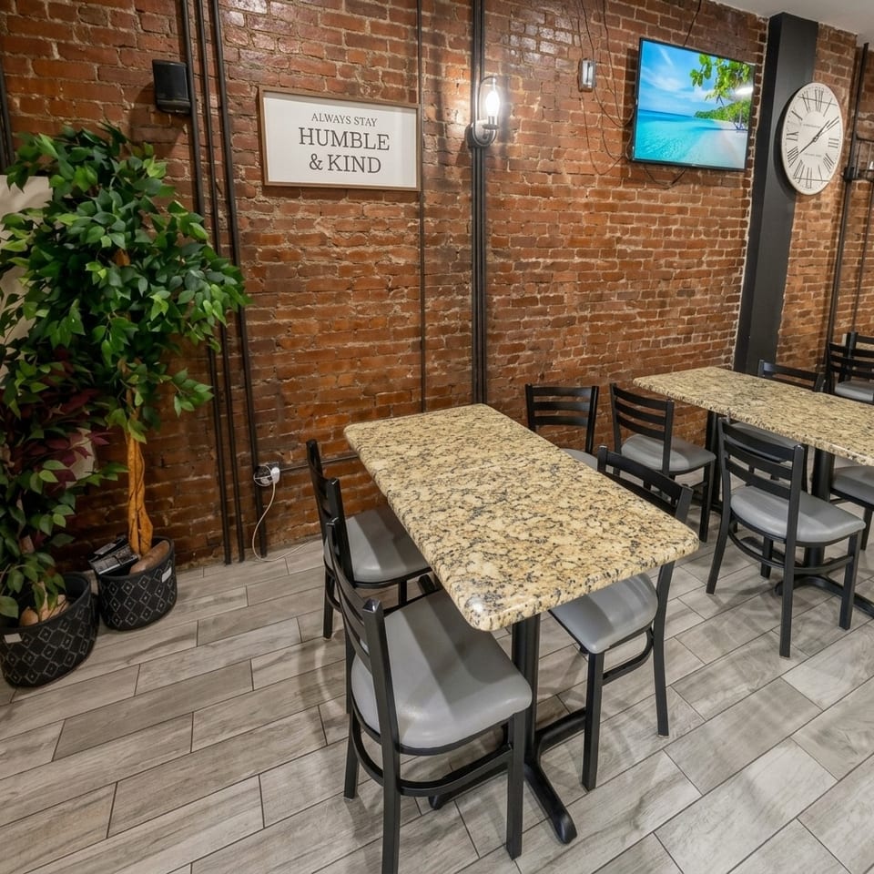 Restaurant dining area with granite tables, black chairs, exposed brick walls, plants, wall art, television, and large clock.