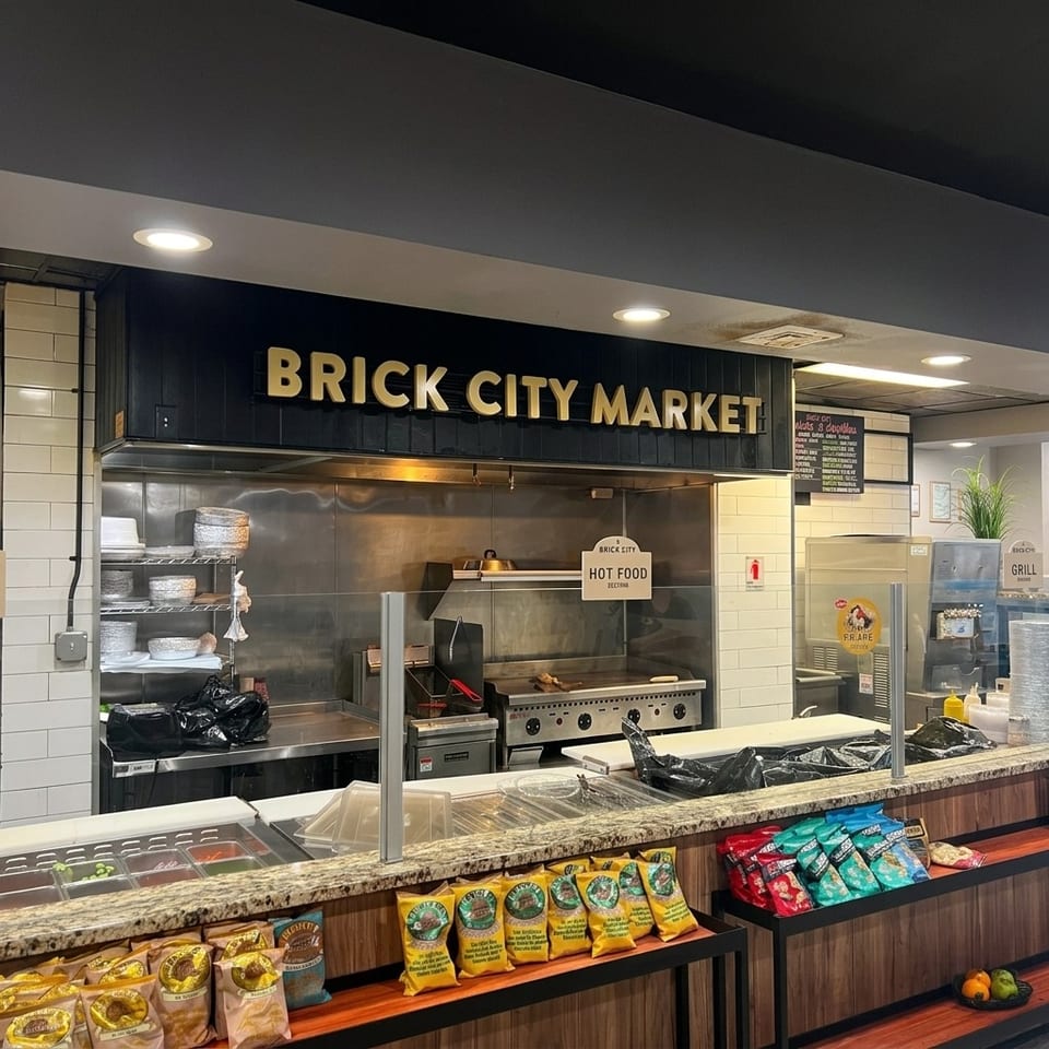 Brick City Market hot-food counter with commercial kitchen equipment, ingredient bins, packaged snacks, and refrigerated drink cases.