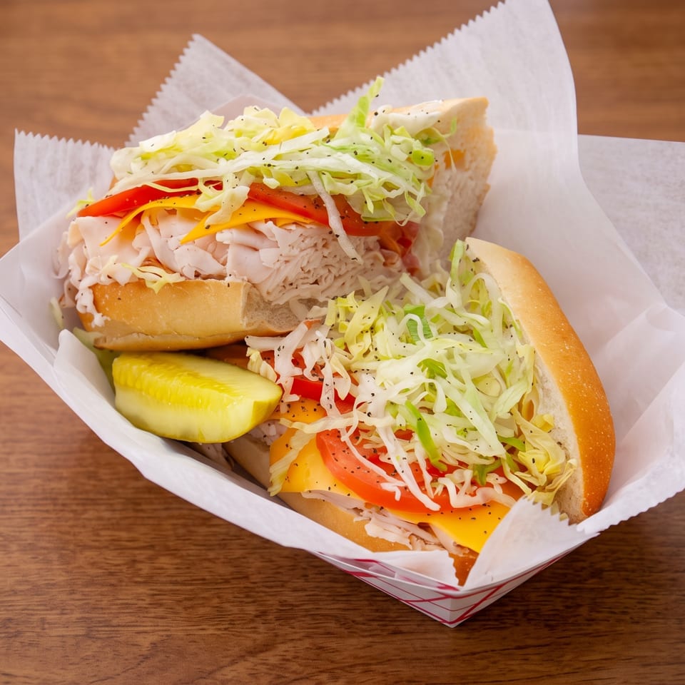 Two chicken sandwiches with lettuce, tomato, cheese, and onions served with a pickle in a paper-lined basket.